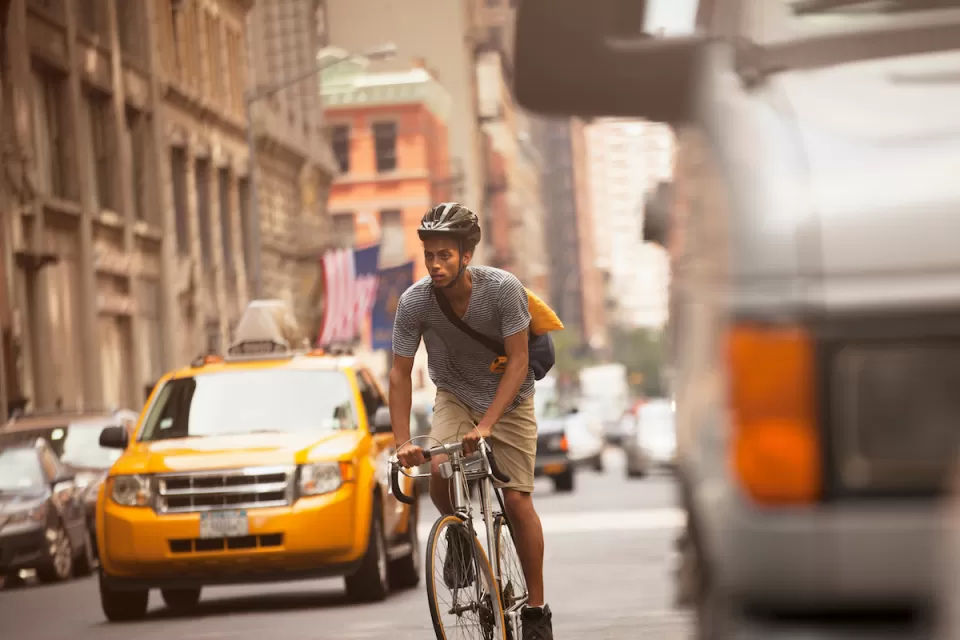 Sepia-toned photo of man riding bicycle on urban street with yellow cab and historic brick buildings in background.